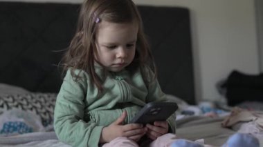 Küçük beyaz bir kız çocuğu akıllı telefonu tutarken evde ağır çekim teknolojisi ve büyüyen kavram olan gerçek insanlar yatağında oturuyor.