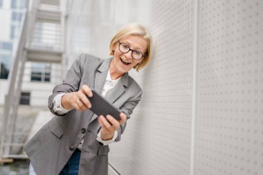 Olgun, sarışın, beyaz bir kadın açık havada oturup mutlu bir gülümseme alıyor video oyunları oynuyor, eğleniyor cep telefonu kullanıyor, akıllı telefon kullanıyor, internet üzerinden gözlük takıyor.