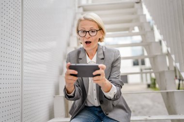 Olgun, sarışın, beyaz bir kadın açık havada oturup mutlu bir gülümseme alıyor video oyunları oynuyor, eğleniyor cep telefonu kullanıyor, akıllı telefon kullanıyor, internet üzerinden gözlük takıyor.