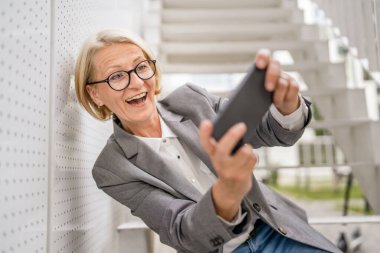 Olgun, sarışın, beyaz bir kadın açık havada oturup mutlu bir gülümseme alıyor video oyunları oynuyor, eğleniyor cep telefonu kullanıyor, akıllı telefon kullanıyor, internet üzerinden gözlük takıyor.