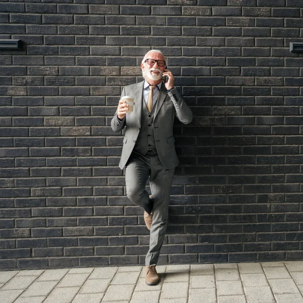 stock image one senior man businessman wear suit with gray beard stand outdoor in front of brick wall of the company hold smartphone and coffee entrepreneur or executive copy space