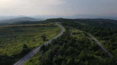 Sırbistan 'da yeşillik ve manzara taşımacılığı ve seyahat konseptiyle çevrili dolambaçlı yol boyunca giden yaz aracı otomobili sırasında bir dağ sırasındaki eğimli asfalt bir yolun hava aracı görüntüleri