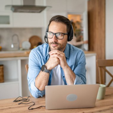 Kafkasyalı bir erkek, evinde kulaklıkla bilgisayar başında çalışıyor. Müşteri destekliyor. Karısının arka planda olduğunu düşünüyor.