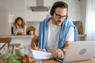 Kafkasyalı bir erkek, evinde kulaklıkla bilgisayar başında çalışıyor. Müşteri destekliyor. Karısının arka planda olduğunu düşünüyor.