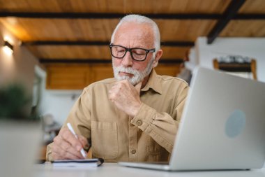 Sakallı ve gözlüklü yaşlı beyaz bir adam dizüstü bilgisayarla çalışıyor. Sorunları çözmek için evde bilgisayar kullanıyor.