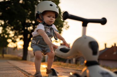 Small caucasian girl toddler playing outdoor in summer evening in sunset with 3 wheel children's kick scooter kid wear protective helmet real people copy space leisure family growing up concept