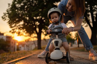Small caucasian girl toddler with mother playing outdoor in summer evening in sunset with 3 wheel children's kick scooter kid wear protective helmet real people copy space leisure family growing up