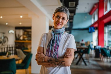 Kafeteryada ya da restoranda tek başına duran, beyaz gömlekli, kendine güvenen, mutlu, mutlu gülümseyen, kısa gri saçlı, modern görünümlü bir kadın.