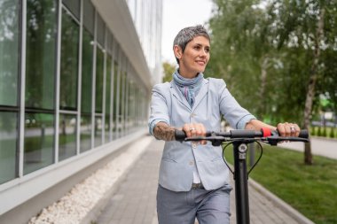 Yetişkin bir son sınıf beyaz kadın elektrik tekmeli scooter 'a biniyor. Şehirde ya da şehirde araba sürüyor. E-scooter çevre dostu.