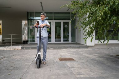 Yetişkin, yaşlı, beyaz bir kadın. Elektrikli scooterın üzerinde duruyor. Şehirde ya da şehirde araba sürüyor.