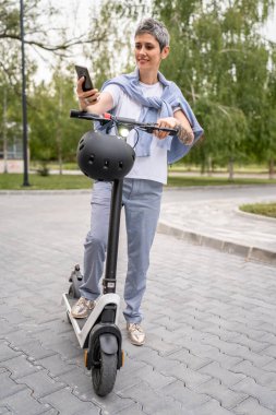 Yetişkin bir son sınıf beyaz kadın dışarıda duruyor selfie çekimleri için cep telefonu kullanıyor ya da elektrik tekmeli scooter ile güveni olan modern alternatif ulaşım aracı fotokopi alanı modunda video çekiyor.