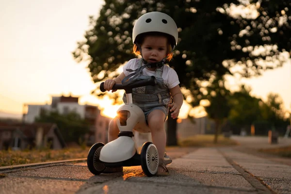 小さな白人の女の子幼児遊び屋外で夏の夜に日没で3輪子供キックスクーター子供は保護ヘルメットを着用リアル人々コピースペースレジャー家族成長コンセプト — ストック写真