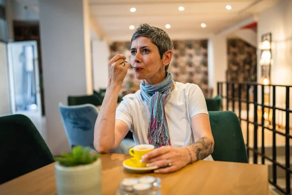 一位年长的高加索女人坐在咖啡店或餐馆里喝着一杯咖啡 真正的人模仿现代灰白短发人独自坐着的空间 — 图库照片