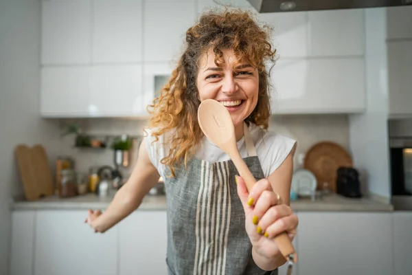 一个快乐的年轻的高加索女人穿着围裙在厨房里笑着拿着混合勺子唱歌 — 图库照片