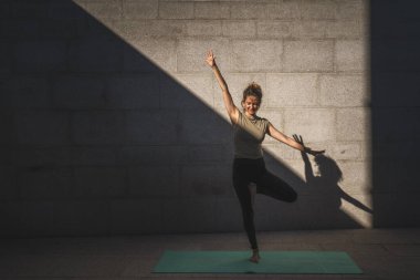 Yetişkin bir Kafkasyalı kadın yaz günü yoga pratiği yapıyor. Şehrin dışında, duvarın önünde. Gölge ve güneş gibi sağlıklı bir yaşam tarzı var.