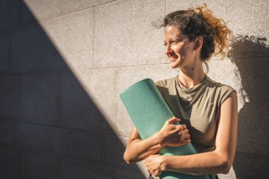 Beyaz bir kadın, güneşli bir günde dışarıda dikilirken yoga minderi tutuyor.