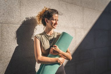 Beyaz bir kadın, güneşli bir günde dışarıda dikilirken yoga minderi tutuyor.