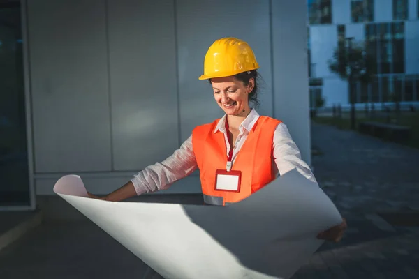 architect woman female construction engineer stand outdoors wear protective helmet and west in front of modern building wall hold construction plans