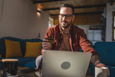 Yetişkin bir beyaz erkek evde dizüstü bilgisayarla mutlu bir gülümseme içinde banka kredi kartı alışverişi yapıyor gerçek insanlar e-ticaret e-bankacılık hizmetlerini kopyalıyor.