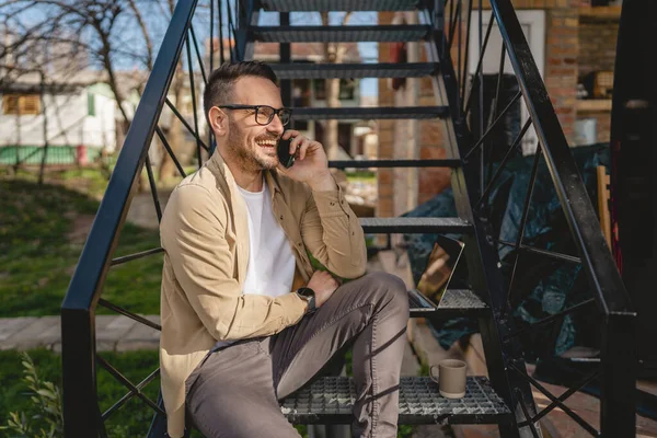 一名男子坐在室外的台阶上 一边用手提电脑刹车一边用手机打电话 一边开心地笑着 自信地笑着 — 图库照片