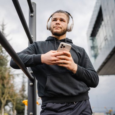 Beyaz tenli genç bir erkek açık hava eğitim parkı spor salonunda duruyor. Kulaklıklı cep telefonu kullanıyor. Gerçek insanları eğitmek için internetten mesajlar yolluyor.