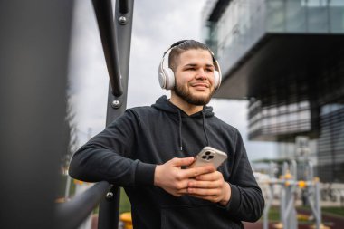 Beyaz tenli genç bir erkek açık hava eğitim parkı spor salonunda duruyor. Kulaklıklı cep telefonu kullanıyor. Gerçek insanları eğitmek için internetten mesajlar yolluyor.