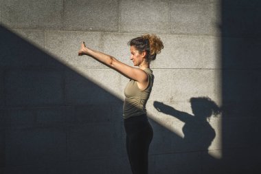 Yetişkin bir Kafkas kadın yaz günü yoga antrenmanı yapıyor şehrin dışında, duvarın önünde gölgesi ve güneşi olan sağlıklı yaşam tarzı konseptiyle kendi başının çaresine bakıyor.