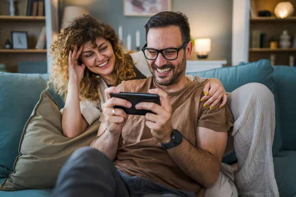 Adult couple man and woman caucasian husband and wife or boyfriend and girlfriend he play video games at home hold mobile phone smart phone have fun leisure joy and bonding concept copy space