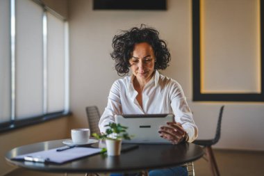 Bir kadın işkadını kafeteryada ya da restoran yöneticisinin masasında oturuyor. İş yerinde dijital tablet kullanıyor. İş yapan yetişkin bir kadın girişimci.