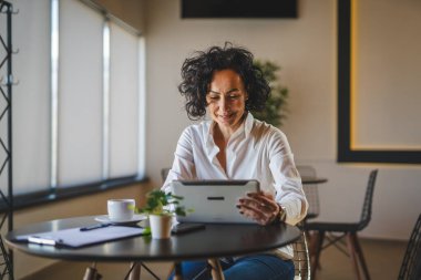 Bir kadın işkadını kafeteryada ya da restoran yöneticisinin masasında oturuyor. İş yerinde dijital tablet kullanıyor. İş yapan yetişkin bir kadın girişimci.