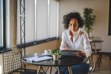 Bir kadın işkadını kafeteryada ya da restoran yöneticisinin masasında oturuyor. İş yerinde dijital tablet kullanıyor. İş yapan yetişkin bir kadın girişimci.