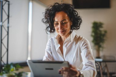 Bir kadın işkadını kafeteryada ya da restoran yöneticisinin masasında oturuyor. İş yerinde dijital tablet kullanıyor. İş yapan yetişkin bir kadın girişimci.