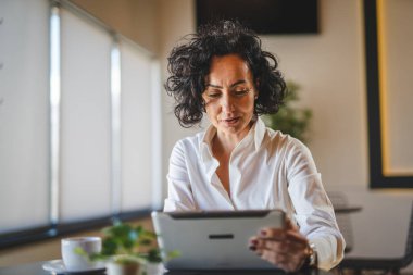 Bir kadın işkadını kafeteryada ya da restoran yöneticisinin masasında oturuyor. İş yerinde dijital tablet kullanıyor. İş yapan yetişkin bir kadın girişimci.