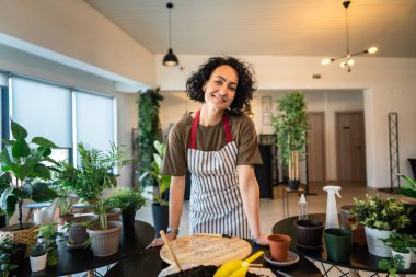 Yetişkin bir kadın elinde çiçek bitkileriyle evde duruyor mutlu gülüşler, ön tarafta belime gülücükler ve botanik bakım konsepti.