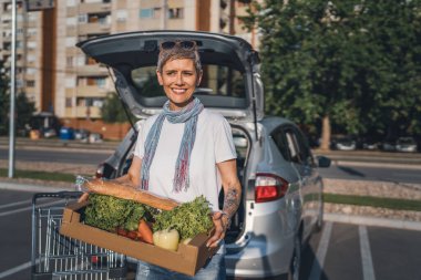 Yetişkin bir kadın, otoparkta duran beyaz bir kadın süpermarketin önünde elinde yiyecek ve sebzelerle duruyor. Mutlu bir gülümseme, sağlıklı vegan konsepti yiyor.