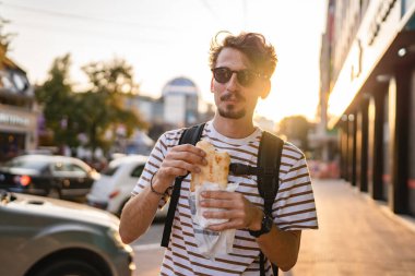 Gündüz vakti şehirde genç, yetişkin, beyaz bir erkek sandviç yiyor. Fast food konsepti. Şehir hayatı.