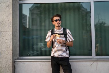 Genç, yetişkin, beyaz bir erkek, güneşli bir günde şehirde duruyor ve sandviç yiyor. Fast food konsepti. Şehir hayatı.