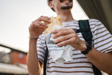 Gündüz vakti şehirde genç, yetişkin, beyaz bir erkek sandviç yiyor. Fast food konsepti. Şehir hayatı.