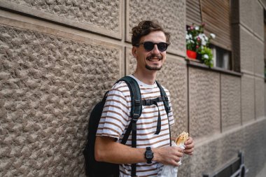 Genç, yetişkin, beyaz bir erkek, güneşli bir günde şehirde duruyor ve sandviç yiyor. Fast food konsepti. Şehir hayatı.