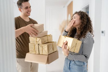 Erkek arkadaşı ya da kocası mutlu bir şekilde gülümserken elinde hediye kutusuyla evde dikilen beyaz bir kadın. Gerçek insanlar sürpriz konsepti kutluyor.