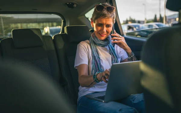ラップトップコンピュータで作業している車の後部座席に座っている1人の成熟した女性白人女性は カジュアルコピースペースを身に着けている道路上の短い灰色の髪の現代と夏の日に電話で話をする — ストック写真