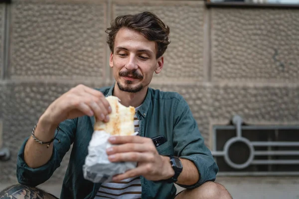 在阳光明媚的日子里 一个年轻的成年男子在城市里站着吃三明治快餐概念城市生活模仿太空游客在城墙前吃饭真实的人 — 图库照片