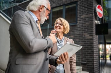 senior man dressed in a suit and a mature blonde woman as business partners husband and wife friends or colleagues talk With digital tablet in hand browse internet look data exchange ideas copy space