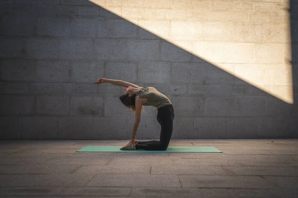 One adult caucasian woman doing yoga practice in summer day outdoor in the city in front of the wall with shadow and sun healthy lifestyle wellbeing concept wide angle full length self care