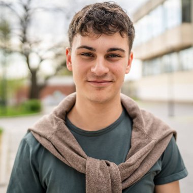 Genç, yetişkin, beyaz bir genç, dışarıda kameraya bakarak poz veriyor. Mutlu, kendine güvenen, sıradan bir gömlek giyiyor.