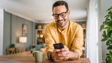 Sakallı ve gözlüklü yetişkin bir adam mutlu bir şekilde gülümseyerek ayakta duruyor cep telefonu akıllı telefonuyla mesaj atıyor ya da internette fotokopi çekiyor.