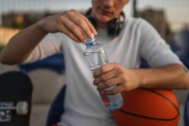 Güneşli bir günde basketbol oynarken, bilinmeyen beyaz bir gencin ellerinde açık plastik bir şişe su.