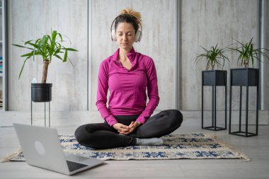Yetişkin bir beyaz kadın milenyum kulaklığı ve dizüstü bilgisayarı kullanarak online meditasyon için bilinçlilik yogası yapıyor. Evdeki gözler kapalı gerçek insanlar mekanı kopyalıyor.