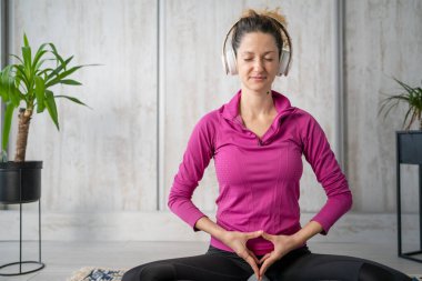 Yetişkin bir beyaz kadın milenyum kulaklığı ve dizüstü bilgisayarı kullanarak online meditasyon için bilinçlilik yogası yapıyor. Evdeki gözler kapalı gerçek insanlar mekanı kopyalıyor.