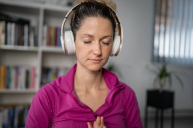 Yetişkin bir beyaz kadın milenyum kulaklıklarını internet rehberli meditasyon için kullanıyor. Evde gözleri kapalı, farkındalık yogası yapıyor. Gerçek insanlar kendi kendilerine bakıyorlar.